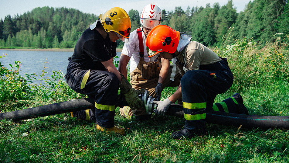 Kaksi palokuntanuorta ja yksi nuorten kouluttaja yhdistävät letkuja järven rannalla.