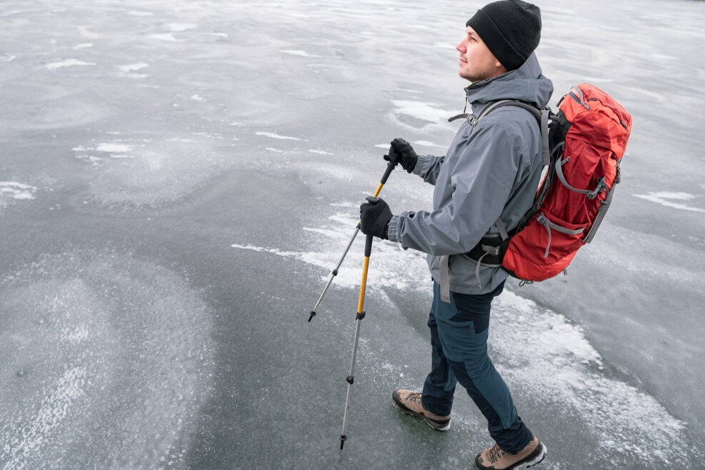 Nuori aikuinen kävelee järven tai meren jäällä. Hänellä on kävelysauvat käsissään ja pieni rinkka selässä.