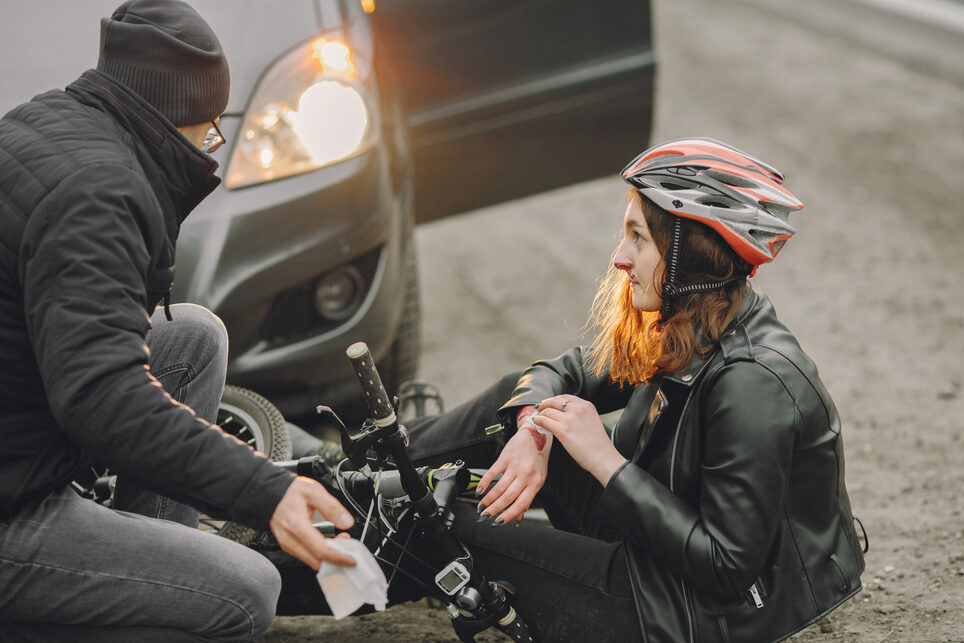 Pyöräilykypäräpäinen nainen istuu asfaltilla pyöränsä vieressä ja pitelee toista rannetta. Häneltä tulee verta nenästä. Vieressä on auto, josta on tullut henkilö auttamaan häntä.