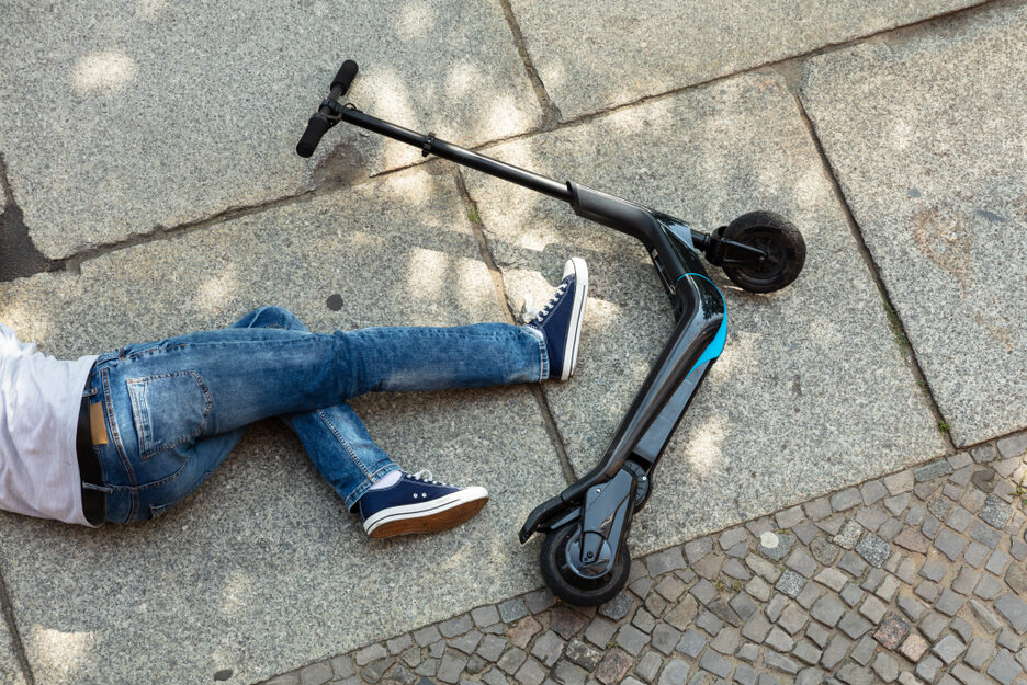 Sähköpotkulauta on maassa katukivetyksellä poikittain ja sen vieressä makaa maassa farkkuihin ja lenkkareihin pukeutunut henkilö, josta näkyy vain alaruumis.