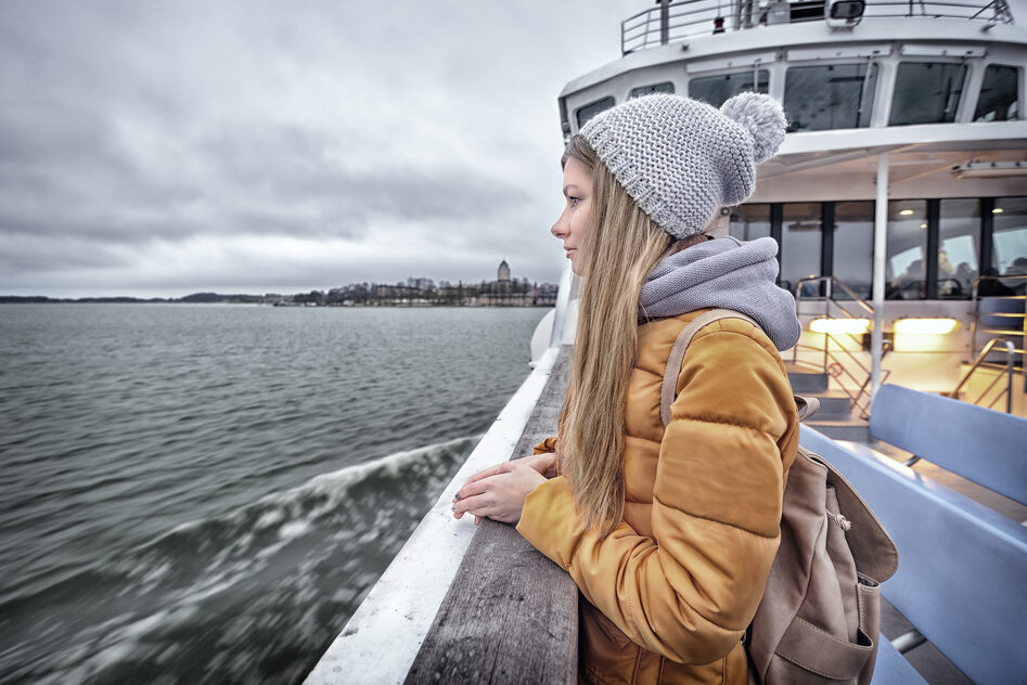 Nuori pipopäinen nainen seisoo Suomenlinnan lautan kannella ja katsoo merelle.