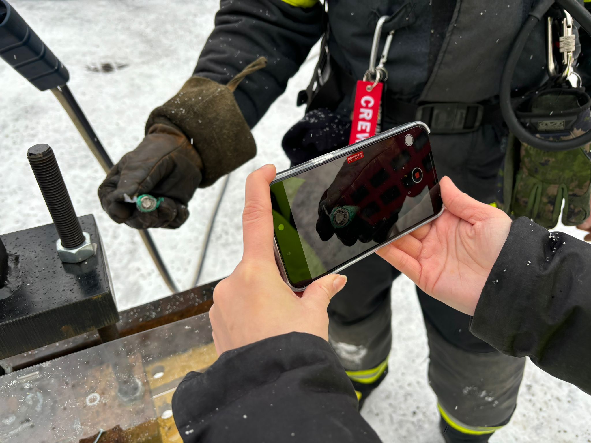 Suojakäsineisiin pukeutunut henkilö pitelee vioittunutta paristoa ja toinen henkilö kuvaa puhelimella.