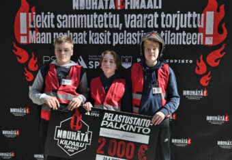 Voittajajoukkue eli kolme kasiluokkalaista poikaa seisoo rivissä kuvausseinällä ja pitää yhdessä kiinni suuresta palkintosekistä.