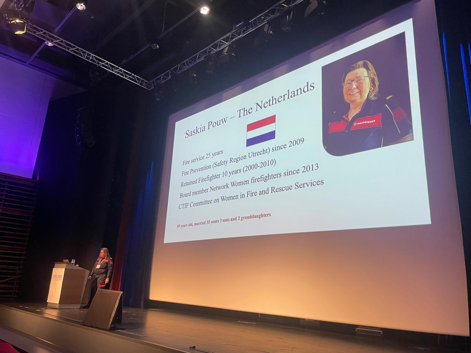 Nainen seisoo isolla esiintymislavalla, jonka valkokankaalla näkyy hänen kuvansa ja tiivistys työhistoriasta.